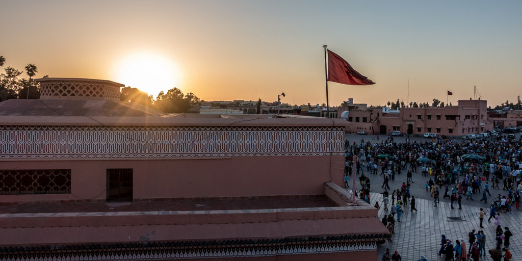 Über einem Marktplatz in Marrakesch geht die Sonne aus. zu sehen ist ein altes Gebäude mit einer wehenden marokksnischen Flagge und eine große Gruppe von Menschen auf dem Marktplatz.