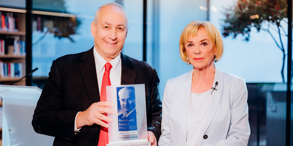 Liz Mohn and Chemi Peres are standing behind a table at the award ceremony for the Reinhard Mohn Prize 2020 and are looking into the camera. Chemi Peres is holding the trophy for the Reinhard Mohn Prize.
