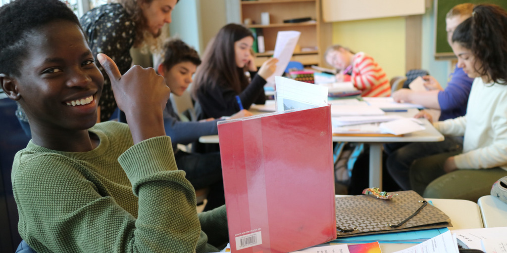 Ein Schüler sitzt an einem Tisch in einem Klassenzimmer, lächelt in die Kamera und macht "den Daumen hoch". Vor ihm steht ein aufgeschlagenes Buch auf dem Tisch. Im Hintergrund sitzen fünf weitere Schüler an einem Tisch und lesen. Über den Tisch gebeut steht eine Lehrerin.