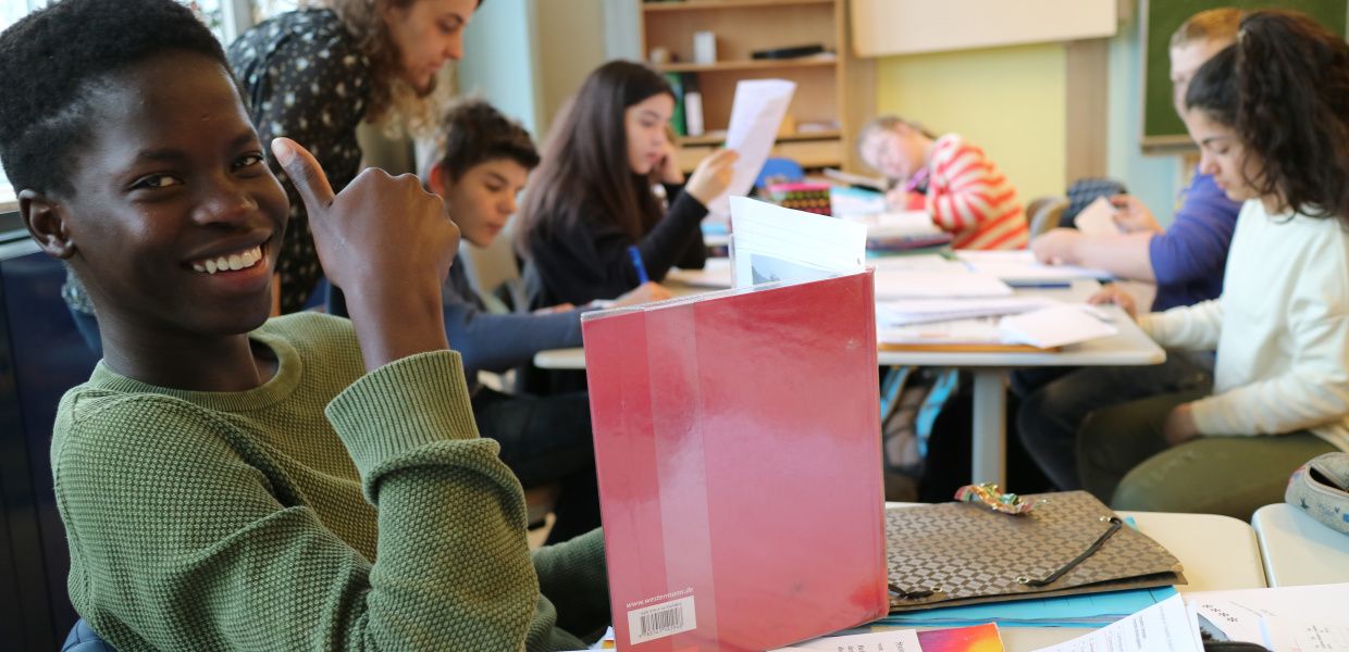 (© Ulfert Engelkes) Ein Schüler sitzt an einem Tisch in einem Klassenzimmer, lächelt in die Kamera und macht "den Daumen hoch". Vor ihm steht ein aufgeschlagenes Buch auf dem Tisch. Im Hintergrund sitzen fünf weitere Schüler an einem Tisch und lesen. Über den Tisch gebeut steht eine Lehrerin.