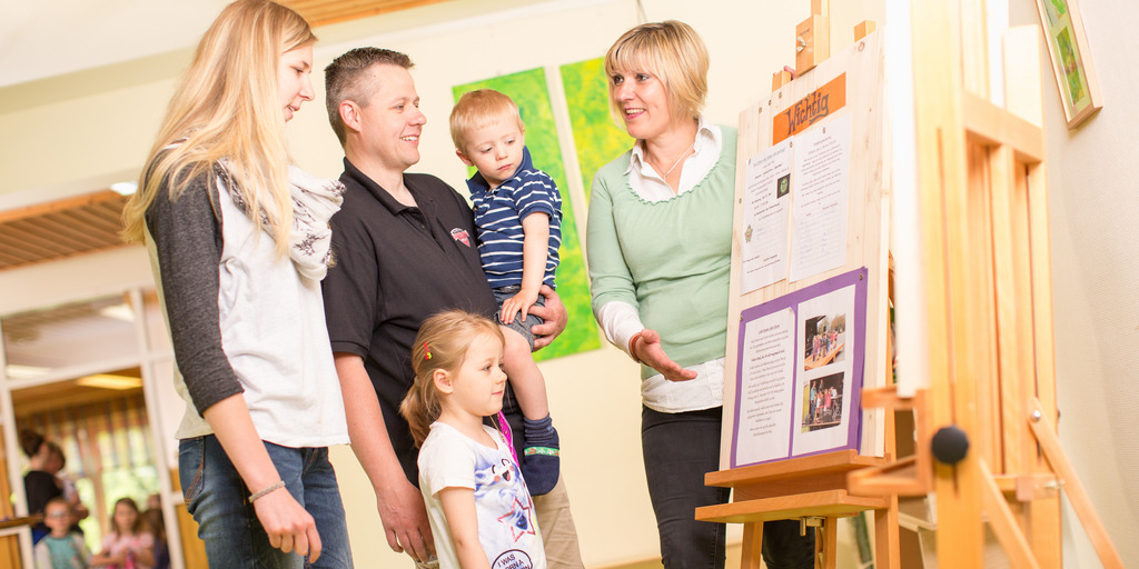 Eine Kita-Leiterin gibt einem Elternpaar mit zwei Kindern an einer Schautafel Informationen zu ihrer Kindertagesstätte.