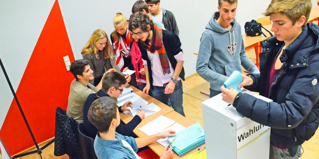 An einer Schule, dem Kolleg St. Blasien in Baden-Württemberg, geben Schüler ihre Stimme bei der Juniorwahl 2013 ab. Ein Schüler wirft seinen Stimmzettel in die Wahlurne, während sich andere Schüler an einem Tisch den Stimmzettel aushändigen lassen.