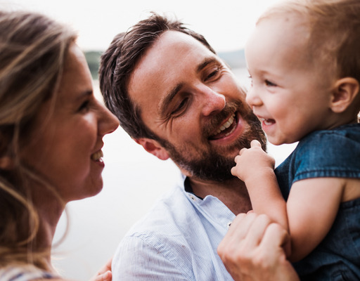 Ein junges Elternpaar mit ihrem kleinen Sohn. Der Vater hat den Sohn auf dem Arm, während die Mutter dem Jungen über den Rücken streichelt. Alle drei lächeln sich gegenseitig an.