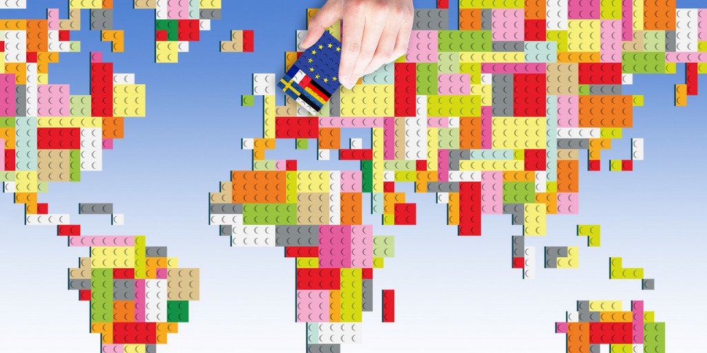 Weltkarte aus Legosteinen dargestellt. Eine Hand hält oberhalb der Karte einen Legostein mit einer EU-, Deutschland, Frankreich, Schweden und Estland Flagge.