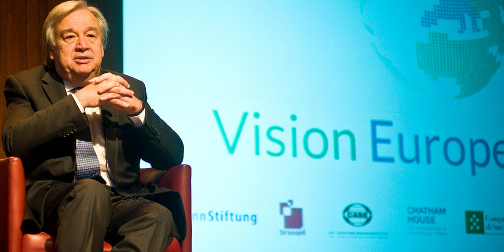 António Guterres speaks on the podium of the Vision Europe Summit while sitting in an armchair. Next to him one can see the Vision Europe Summit logo displayed on a large video screen.
