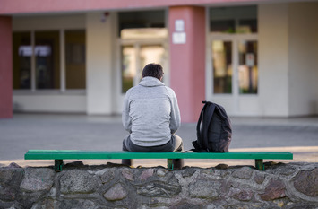 Ein Jugendlicher ist von hinten zu sehen, wie er mit gesenktem Blick alleine auf einer Bank sitzt.