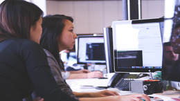 Zwei Frauen sitzen vor zwei Bildschirmen eines Computers. Im Hintergrund sieht man weitere Bildschirme.