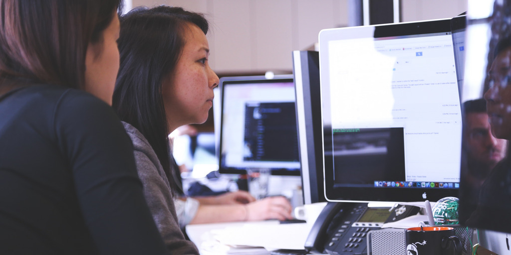 Zwei Frauen sitzen vor zwei Bildschirmen eines Computers. Im Hintergrund sieht man weitere Bildschirme.