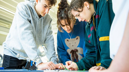 Drei Jugendliche beugen sich im Rahmen eines Lerncamps über einen Tisch und arbeiten gemeinsam an einer kleinen elektronischen Konstruktion.