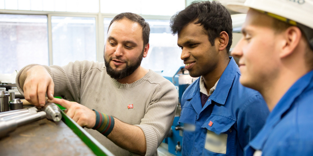 Ein Mitarbeiter zeigt den Azubis, darunter einem Flüchtling, mit der Drehbank gefertigte Werkstücke