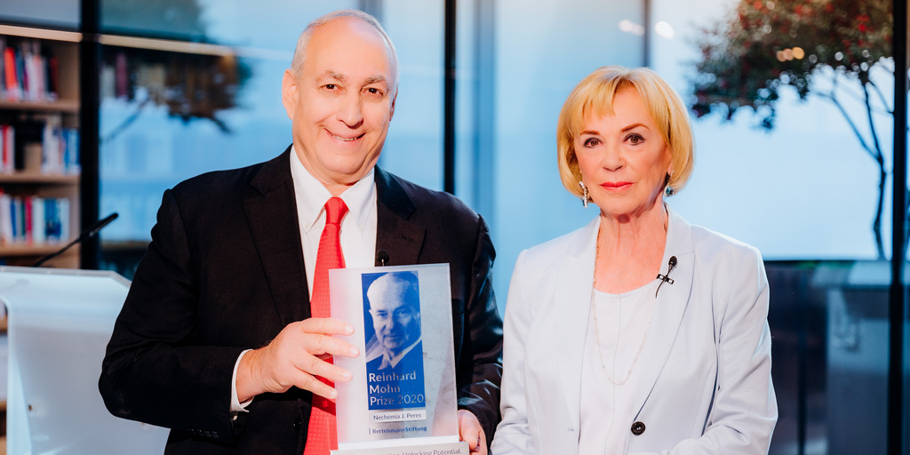 Liz Mohn und Chemi Peres stehen bei der Übergabe des Reinhard Mohn Preises 2020 an einem Tisch und blicken in die Kamera, Chemi Peres hält die Preis-Stele in der Hand.