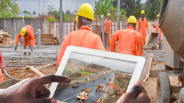 Zwei Hände halten ein Notebook, in dem eine Baustelle zu stehen ist. Im Hintergrund ist ebenfalls eine - scheinbar afrikanische -Baustelle zu sehen.