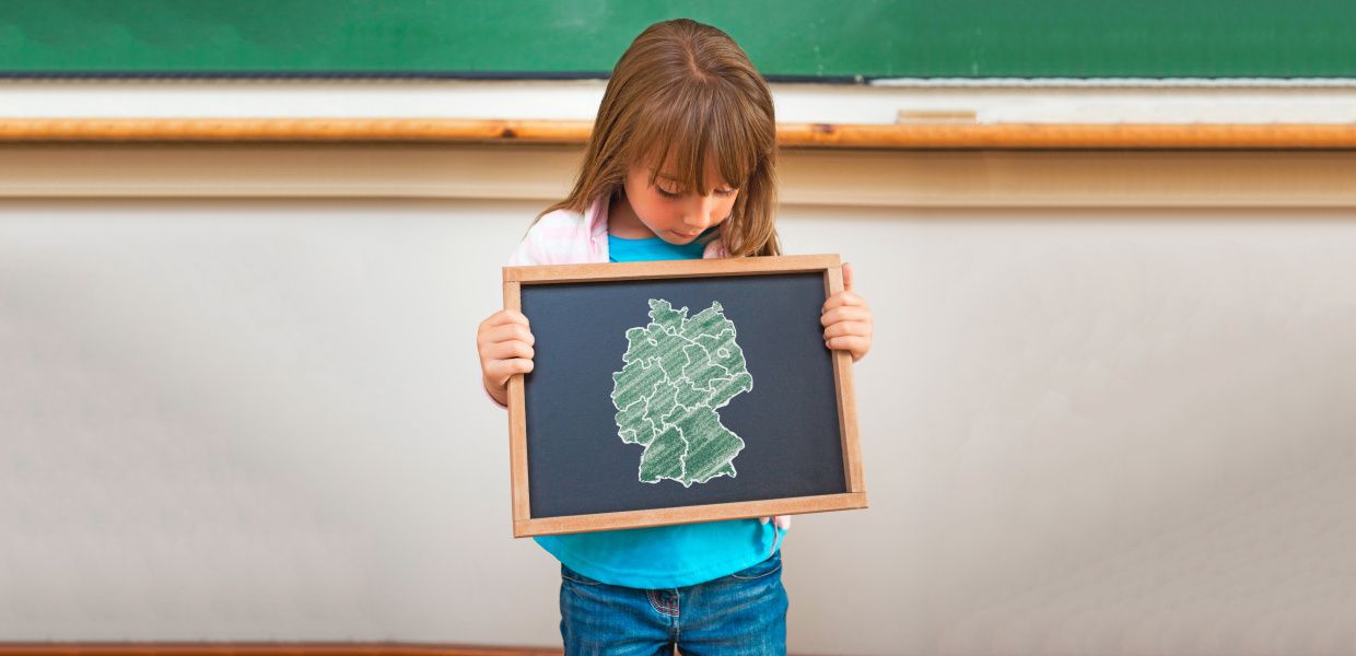 (© Shutterstock/wavebreakmedia; PantherMedia/Stefan Kunert) Ein Mädchen blickt auf eine kleine Tafel, die es in seinen Händen hält und auf der eine Deutschlandkarte mit den Umrissen der 16 Bundesländer aufgezeichnet ist. Im Hintergrund eine große Schultafel.