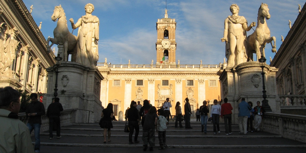Blick von den Treppen, die hinauf zum Kapitolshügel in Rom führen, auf den Senatorenpalast. Links und rechts säumen zwei große Statuen mit Pferden den Weg. Auf der Treppe laufen Menschen.