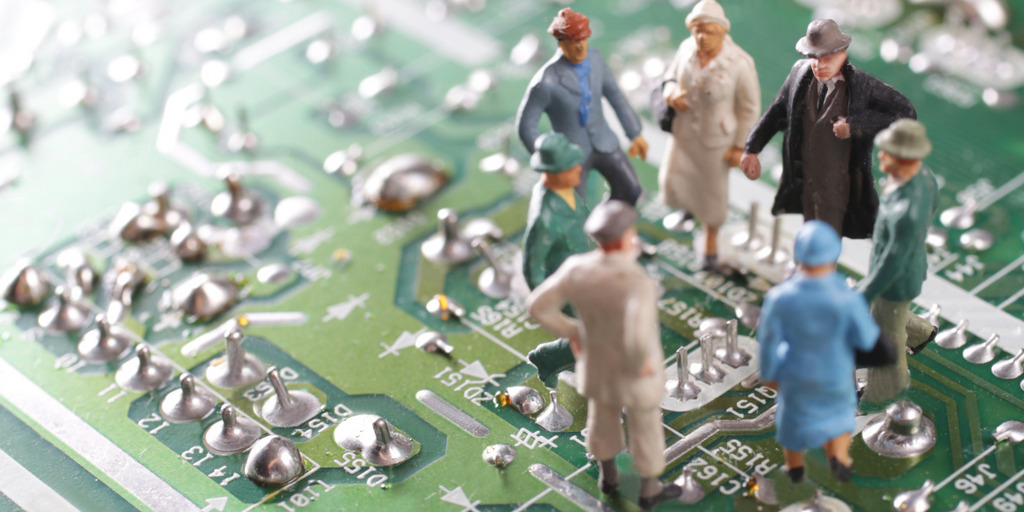 A group of elder people, represented by small figurines, is standing on the printed circuit board of a computer and talking to each other.