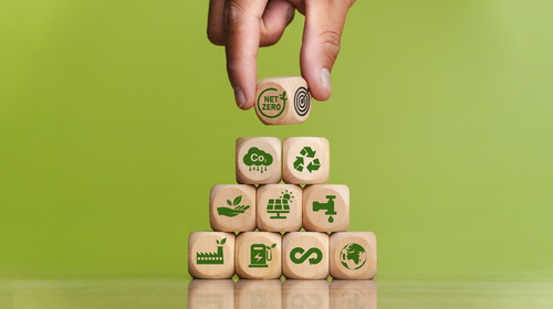 The picture shows wooden cubes stacked to form a pyramid in front of a green wall. Various symbols relating to sustainability can be seen on the cubes.