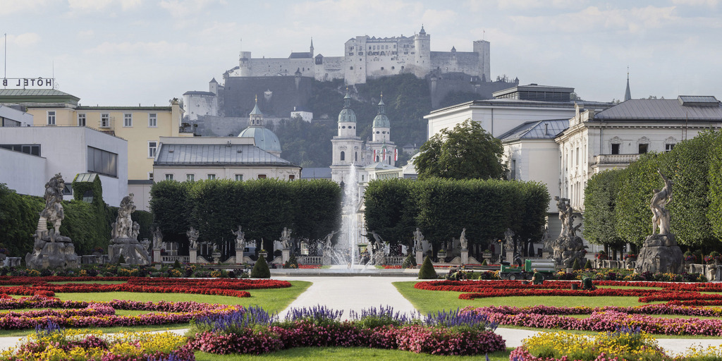 Blick in den Mirabellgarten in Salzburg. Im Vordergrund sieht man in geometrischen Formen angepflanzte Blumenbeete, weiter im Hintergrund eine Fontäne sowie den Salzburger Dom und die Festung Hohensalzburg, die über der Stadt thront. Das Bild wurde kurz vor Beginn des Salzburger Trilogs 2023 aufgenommen.
