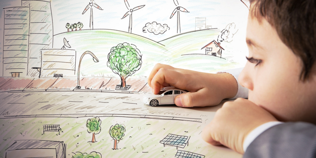 Ein kleiner Junge spielt mit einem Spielzeugauto. Er schiebt es über eine gezeichnete Straße, um die herum Häuser, Bäume und, als Symbol für Nachhaltigkeit, Windräder und Solar-Panels stehen.