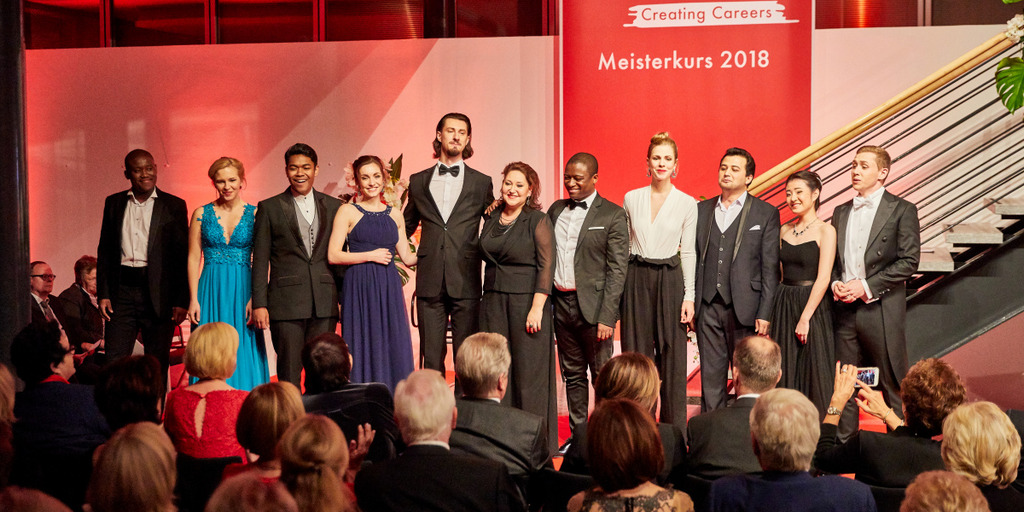 Gruppenbild der Teilnehmerinnen und Teilnehmer des Meisterkurses NEUE STIMMEN nach dem Abschlusskonzert. Sie stehen auf der Bühne im rot beleuchteten und mit Blumen geschmückten Foyer der Bertelsmann Stiftung. Das Publikum applaudiert, in der ersten Reihe NEUE-STIMMEN-Präsidentin Liz Mohn.