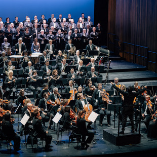 Blick auf die Bühne der Oper Bonn mit dem Orchester und dem Chor. Vor dem Chor sitzen die Solist:innen.