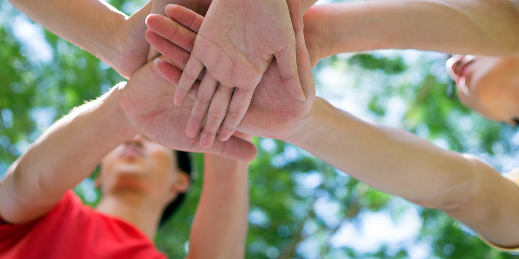 Mehrere Jugendliche stehen im Kreis und legen in der Mitte des Kreises ihre Hände aufeinander, um Verbundenheit und ein Teamgefühl auszudrücken.