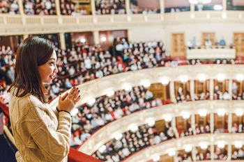 Eine junge Frau steht an einer Balustrade in einem vollbesetzten Theater oder Opernhaus und applaudiert.