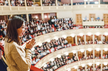 Eine junge Frau steht an einer Balustrade in einem vollbesetzten Theater oder Opernhaus und applaudiert.