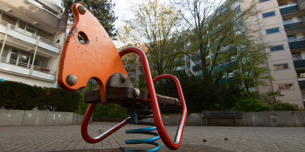 Eine Kinderwippe in Form eines Pferdekopfs auf einem gepflasterten Platz. Im Hintergrund Bäume und Wohnblöcke.