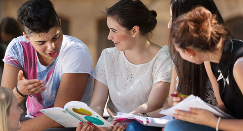 Eine Gruppe Schüler:innen sitzt zusammen auf dem Schulhof und schaut gemeinsam in Schulhefte und diskutiert miteinander