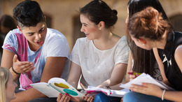 Eine Gruppe Schüler:innen sitzt zusammen auf dem Schulhof und schaut gemeinsam in Schulhefte und diskutiert miteinander