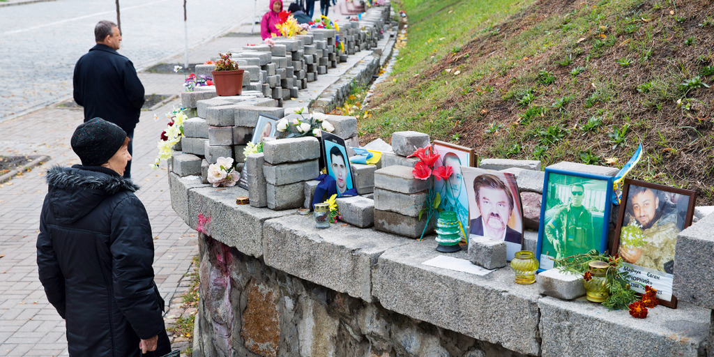 Mauer am Rande des Majdan, dem zentralen Platz der ukrainischen Hauptstadt Kiew. Auf der Mauer stehen zwischen Steinen und Blumen Portraits von bei den Euromajdan-Protesten 2013 und 2014 getöteten Demonstranten. Passanten laufen vorbei und blicken auf di