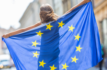 Ein Mädchen breitet mit ausgestreckten Armen hinter seinem Rücken eine Europaflagge aus und läuft so durch eine Stadt.