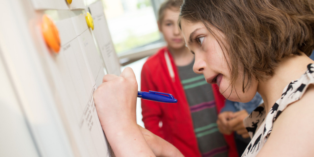 Eine Schülerin macht Notizen in einer an einer Magnetwand hängenden Tabelle. Dabei sieht ihr ein im Hintergrund stehender Mitschüler zu.