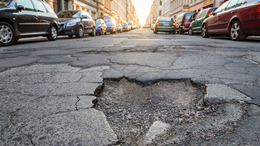Das Bild zeigt eine innerstädtische Straße mit parkenden Autos rechts und links, die deutliche Fahrbahnschäden aufweist. Im Vordergrund ist ein großer, tiefer Schlagloch zu sehen, umgeben von rissigem und brüchigem Asphalt.