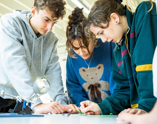 Drei Jugendliche beugen sich im Rahmen eines Lerncamps über einen Tisch und arbeiten gemeinsam an einer kleinen elektronischen Konstruktion.