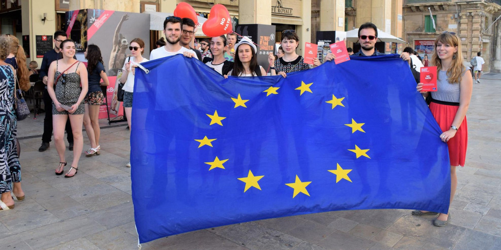 Sieben junge Frauen und Männer stehen auf einem Platz vor einem Gebäude in Malta und halten eine Europafahne in den Händen. Zwei von ihnen halten außerdem Luftballons in Herzform.