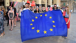 Sieben junge Frauen und Männer stehen auf einem Platz vor einem Gebäude in Malta und halten eine Europafahne in den Händen. Zwei von ihnen halten außerdem Luftballons in Herzform.