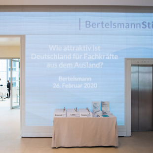Der Check-In-Tisch am Veranstaltungsort. Im Hintergrund ist der Veranstaltungstitel "Wie Attraktiv ist Deutschland für Fachkräfte aus dem Ausland?" an die Wand projiziert
