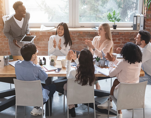 Junge Mitarbeiter:innen eines Start-up-Unternehmens sitzen während einer Besprechung um einen Tisch, lachen und applaudieren.