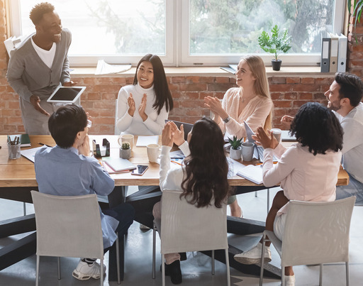 Junge Mitarbeiter:innen eines Start-up-Unternehmens sitzen während einer Besprechung um einen Tisch, lachen und applaudieren.