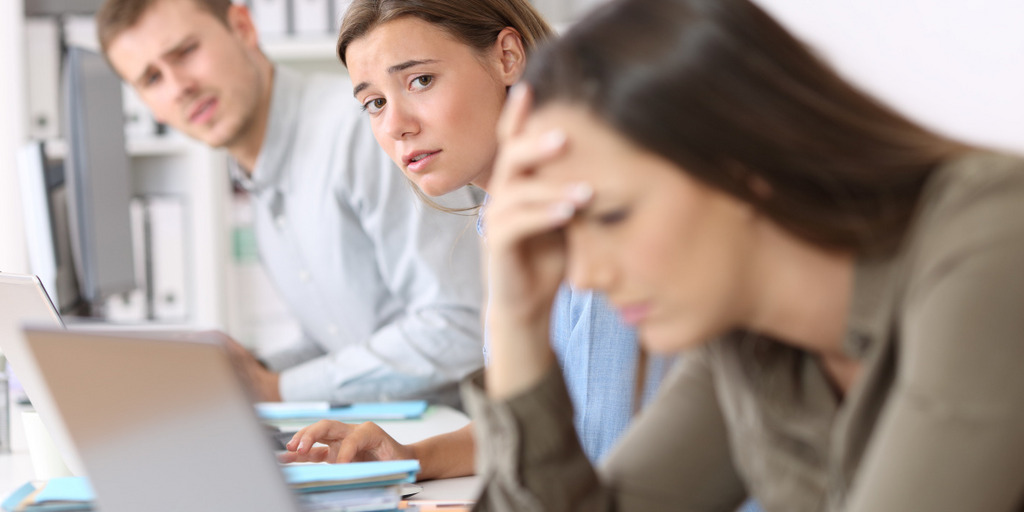 Eine junge Frau blickt angestrengt, mit der Hand an ihrer Stirn und vornüber gebeugt, auf ihren Laptop. Im Hintergrund sehen zwei neben ihr sitzende Kollegen sie mit besorgtem Blick an.