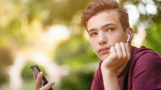 Ein Jugendlicher hat seinen Kopf auf seine linke Hand aufgestützt und blickt nachdenklich in die Ferne, während er in der rechten Hand sein Smartphone hält.