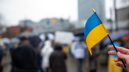 In a big city whose buildings can be seen in the background, a group of people protests against the war in Ukraine. A person in the foreground is holding a small Ukrainian flag into the camera.