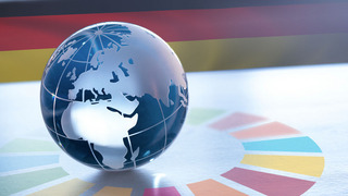 Eine Weltkugel aus Glas liegt auf einem Untergrund, auf dem strahlenförmig von ihm das Logo für die Nachhaltigkeitsziele der Vereinten Nationen ausgeht. Im Hintergrund sieht man die Farben der deutschen Flagge.