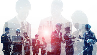 Business people holding laptops and tablets in their hands and engineers wearing construction helmets are shown standing in a row and talking to each other in small groups.