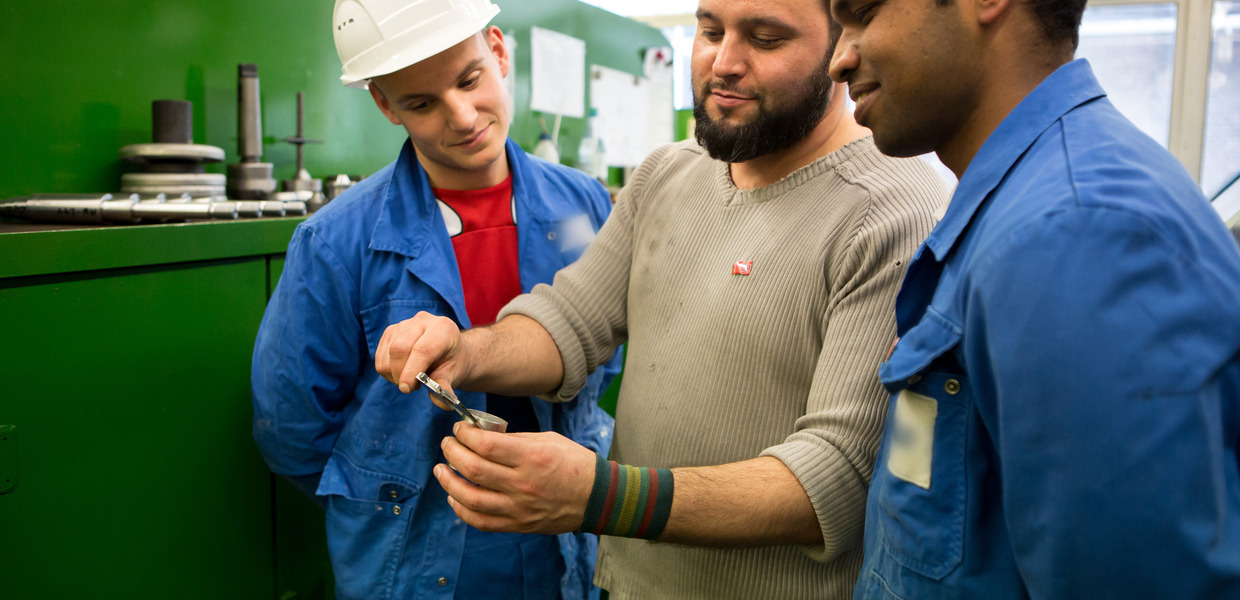 Ein Mitarbeiter zeigt Azubis mit der Drehbank gefertigte Werkstücke.