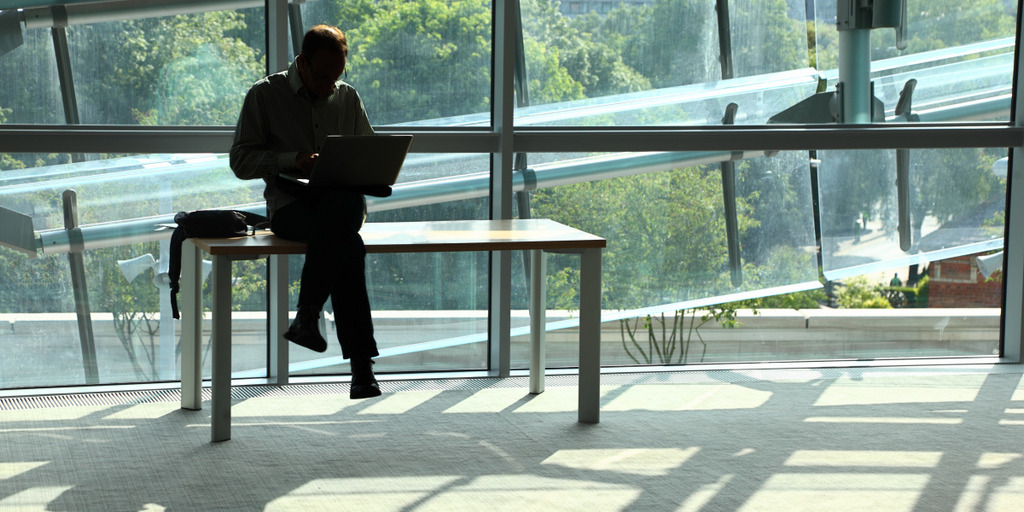 Ein Mann sitzt mit überkreuzten Beinen auf einem Tisch in einem sonnendurchfluteten Raum und arbeitet auf einem Laptop.