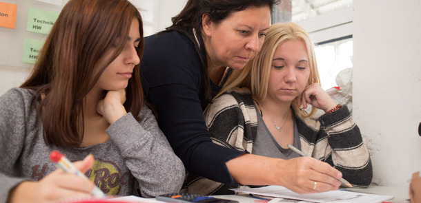 Eine Lehrerin beugt sich von hinten zwischen zwei Schülerinnen durch und korrigiert etwas im Heft der Schülerin