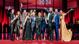 The finalists of NEUE STIMMEN 2024 are posing for a group photo with NEUE STIMMEN founder Liz Mohn and jury chairman Dominique Meyer on the stage of Stadthalle Gütersloh after the semifinal. The singers are cheering.