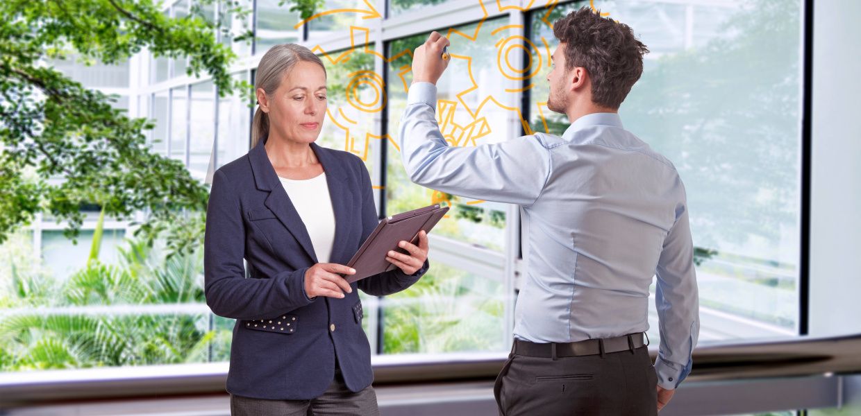 (© Katrin Biller Fotografie, baona / iStockphoto.com, 4zevar / Shutterstock) Eine Unternehmerin sieht auf ihr Tablet, während ein Jungunternehmer neben ihr Zahnräder auf eine Glasscheibe zeichnet.